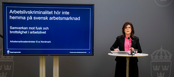 Arbetsmarknadsminister Eva Nordmark står och talar på en pressträff. Bredvid henne finns en stor skärm med en presentation där det står "Arbetslivskriminalitet hör inte hemma på svensk arbetsmarknad".