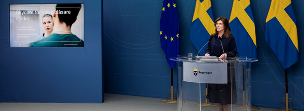 Bild från pressträff i regeringens pressrum. Arbetsmarknadsminister Eva Nordmark står i en talarstol med svenska flaggor och EU-flaggan bakom sig. Bredvid henne syns en presentation där man skymtar ordet visselblåsare.