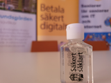 Bild på en liten flaska med texten "Säkert så klart" och i bakgrunden rollup med texten "betala säkert digitalt"