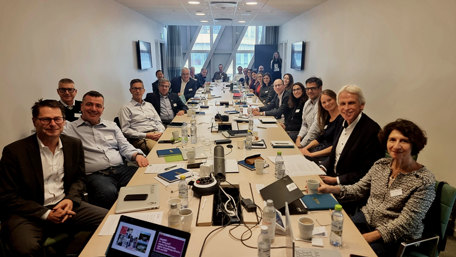 A meeting room with many people, looking into the camera, around a squared table.