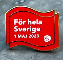 Syntolkning av bild: Första maj märket 2023 som ser ut som två röda fanor där fanan i förgrunden har texten För hela Sverige 1 Maj 2023, samt partiloggan (rosen).